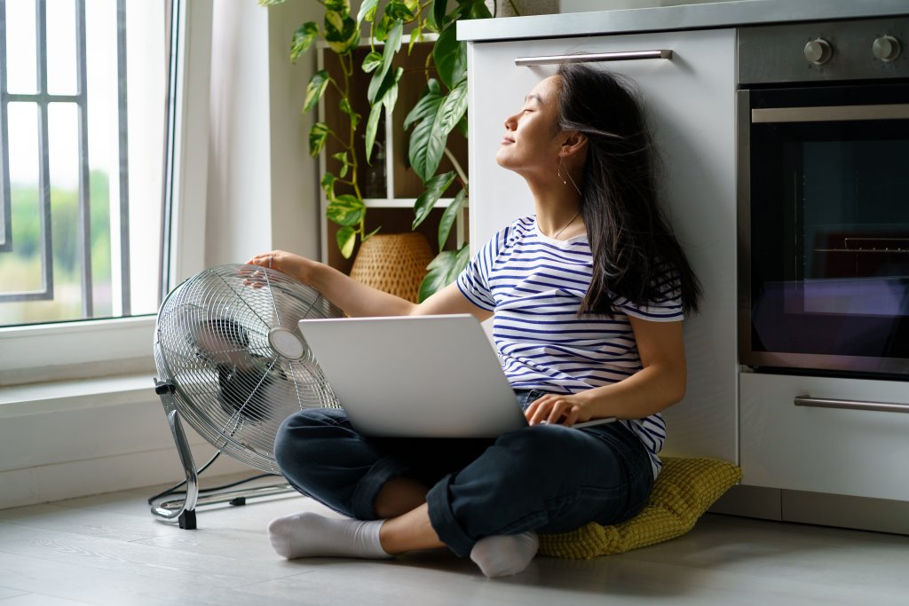 Carefree positive Asian woman working sits in kitchen on floor with laptop and touch at electric fan