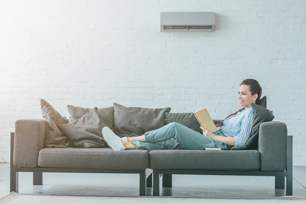happy woman reading book on couch, air conditioner on wall, summer heat